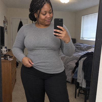 Woman in a fitted grey top at a bedroom mirror, waistline smooth over the CIERRA waist trainer